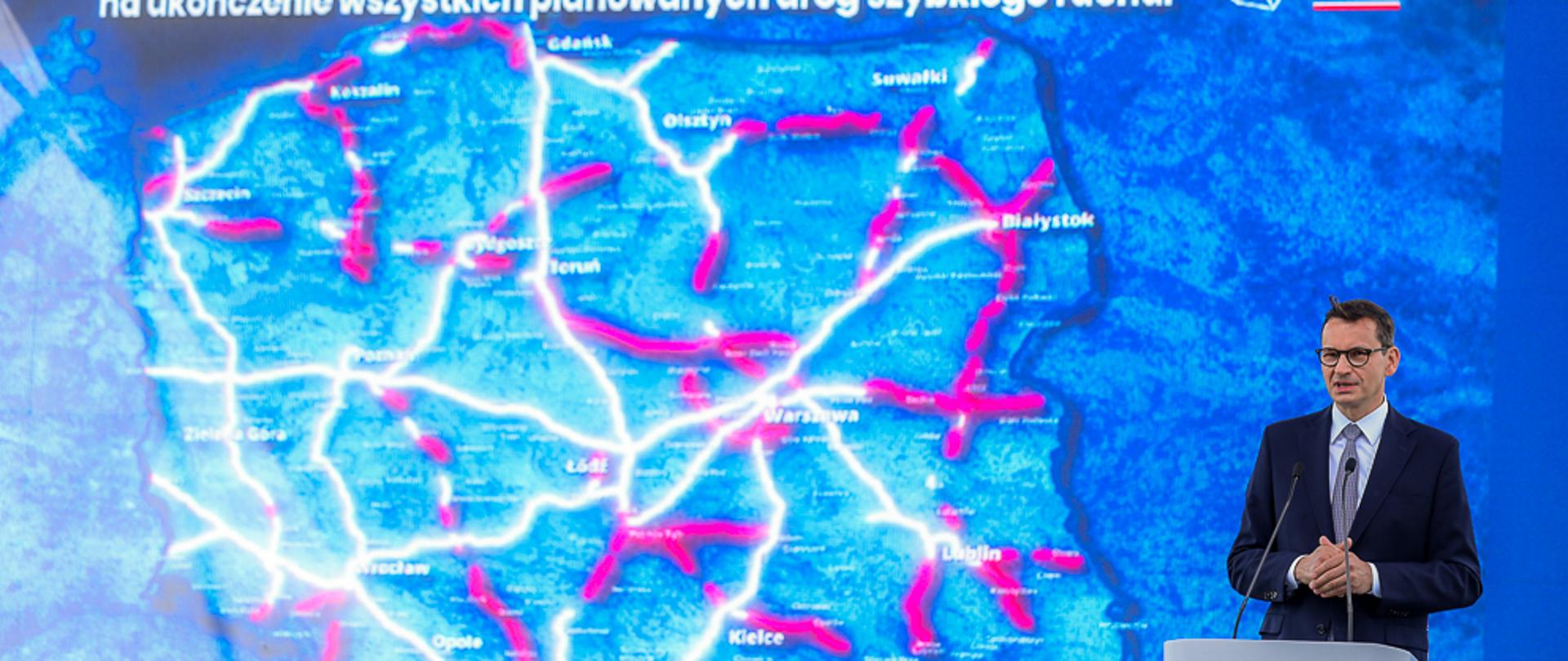 Premier Mateusz Morawiecki w trakcie przemówienia, w tle plansza z mapą kraju z zaznaczonymi odcinkami dróg do modernizacji. Wypowiedź premiera podczas prezentacji Rządowego Programu Budowy Dróg Krajowych 2030.