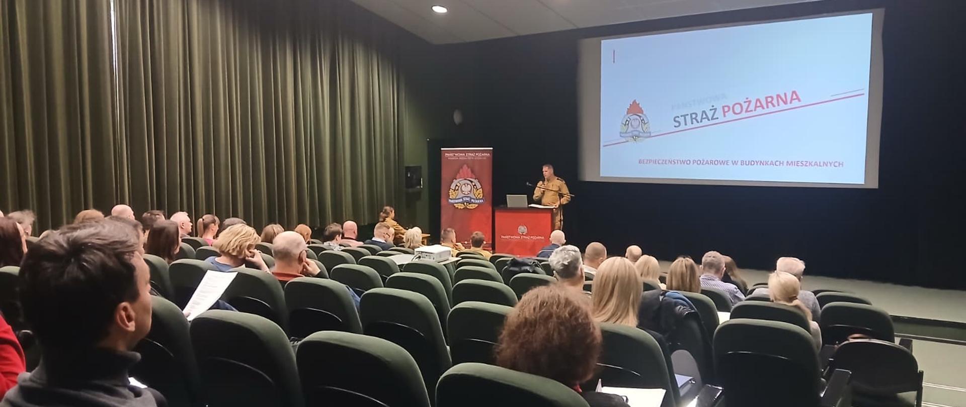 Strażak Państwowej Straży Pożarnej prowadzi prezentację dotyczącą bezpieczeństwa pożarowego w budynkach mieszkalnych podczas spotkania z zarządcami i administratorami nieruchomości w sali kinowej Biblioteki Publicznej w Koszalinie. Na ekranie widoczna jest prezentacja „Straż Pożarna – Bezpieczeństwo pożarowe w budynkach mieszkalnych”, a uczestnicy siedzą na widowni i słuchają wystąpienia.