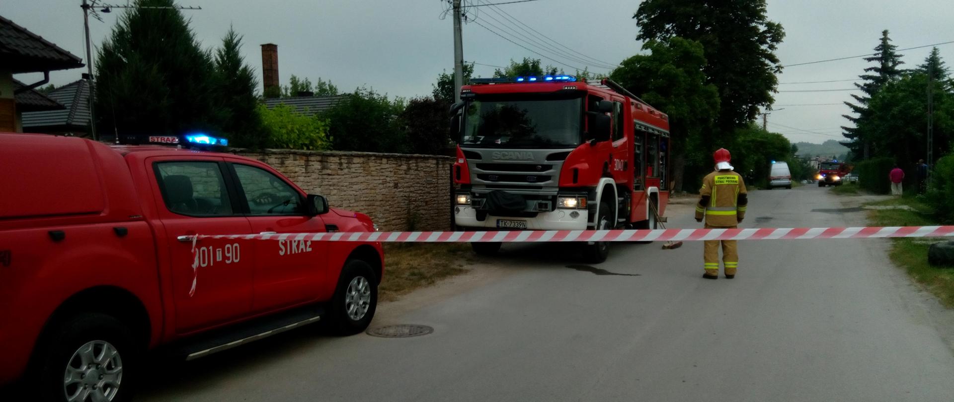 Zdjęcie przedstawia dwa samochody straży pożarnej stojącą wzdłuż ulicy, teren akcji odgrodzony taśma ostrzegawczą oraz strażaka pilnującego wyznaczonej strefy. W tle widać kolejny samochód straży pożarnej oraz pogotowia gazowego.