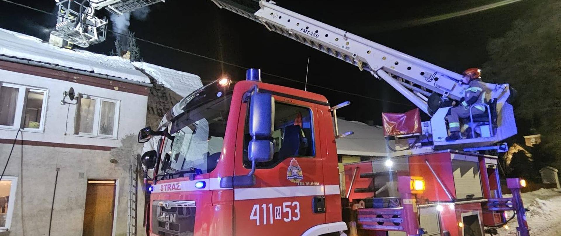 Zdjęcie zrobione nocą. Podnośnik strażacki rozstawiony przy budynku mieszkalnym. Strażacy przy kominie budynku. Dach pokryty śniegiem.