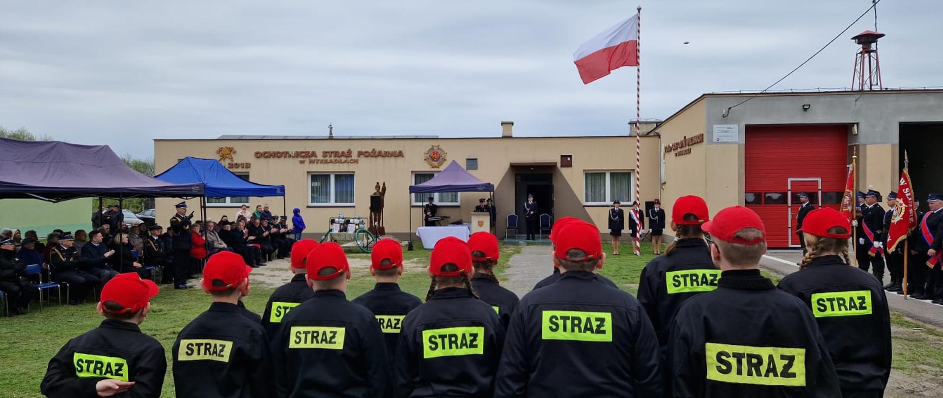 Na pierwszym planie zdjęcia widoczni są strażacy MDP. Na głowach mają czerwone czapki a na plecach żółty napis Straż. Ustawieni są przodem do uroczystości. W tle powiewa flaga Polski na maszcie.