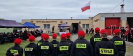 Na pierwszym planie zdjęcia widoczni są strażacy MDP. Na głowach mają czerwone czapki a na plecach żółty napis Straż. Ustawieni są przodem do uroczystości. W tle powiewa flaga Polski na maszcie.