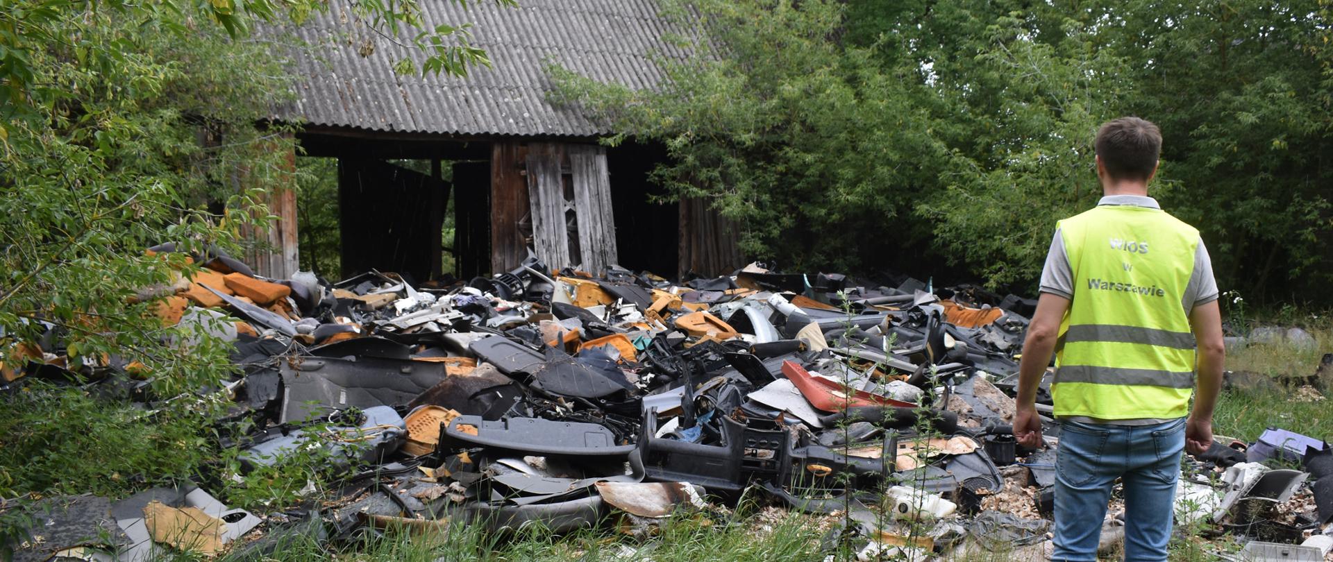 Inspektor Wojewódzkiego Inspektoratu Ochrony Środowiska w Warszawie stoi przed rozrzuconymi na ziemi odpadami z demontażu pojazdów. W tle znajduje się budynek gospodarczy.
