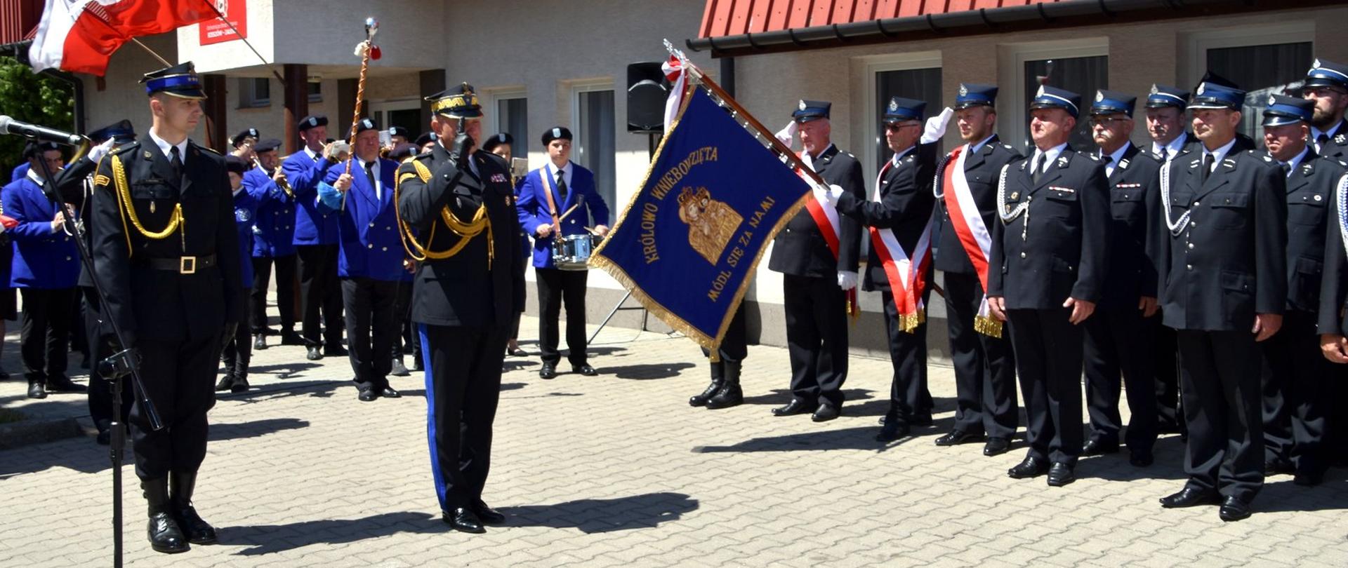 Zdjęcie zrobione na zewnątrz podczas uroczystego apelu. Dwóch oficerów Państwowej Straży Pożarnej stoi na placu, jeden z nich w stopniu generała. Generał salutuje sztandarowi jednostki OSP.