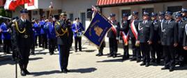 Zdjęcie zrobione na zewnątrz podczas uroczystego apelu. Dwóch oficerów Państwowej Straży Pożarnej stoi na placu, jeden z nich w stopniu generała. Generał salutuje sztandarowi jednostki OSP.