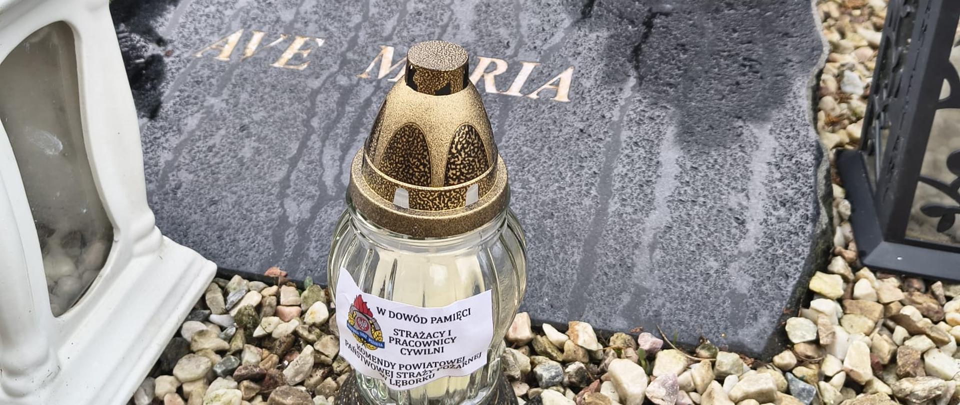 Znicz z pamiątkową naklejką od strażaków stoi na kamiennym nagrobku strażaka.