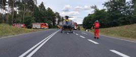 Stojący na jezdni śmigłowiec Lotniczego Pogotowia Ratunkowego. W okolicy widoczne pojazdy pożarnicze oraz umundurowani strażacy. Po prawej stronie w rowie widoczne leżące na boku auto osobowe.