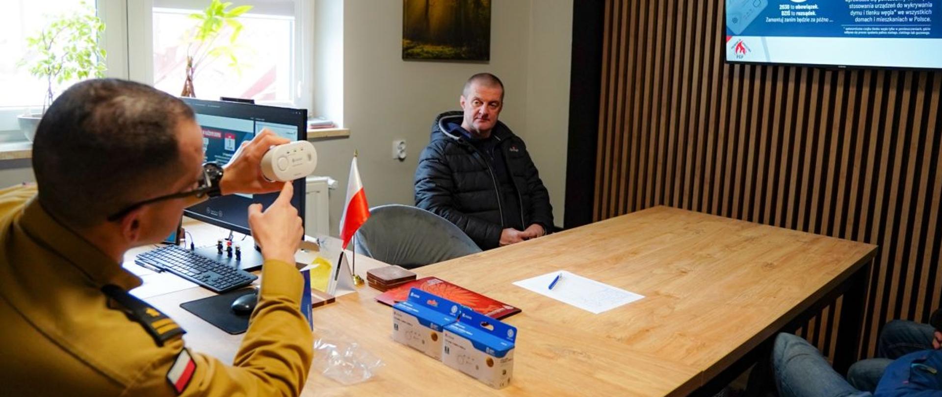 Zdjęcie przedstawia zastępce komendanta z Ustrzyk Dolnych który wyjaśnia zasadę działania czujki dymu mężczyznom siedzącym przy stole. Za nimi na ścianie wisi telewizor na którym widać obraz z czujką dymu i opisem. 