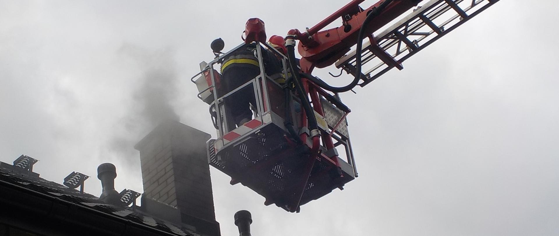 Fotografia przedstawia fragment dachu budynku z widocznym kominem. Przy kominie w koszu podnośnika widać dwóch strażaków w ubraniach bojowych i hełmach podejmujących działania ratowniczo gaśnicze wynikające z zapalenia się sadzy w przewodzie kominowym.
