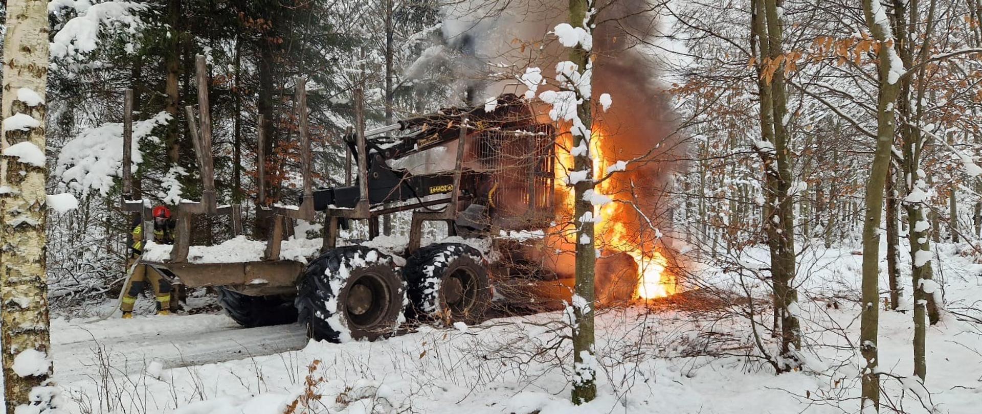 Maszyna leśna Forwarder objęta pożarem. Strażacy ubrani w ubrania ochronne koloru piaskowego i sprzęt ochrony układu oddechowego gaszą pożar.