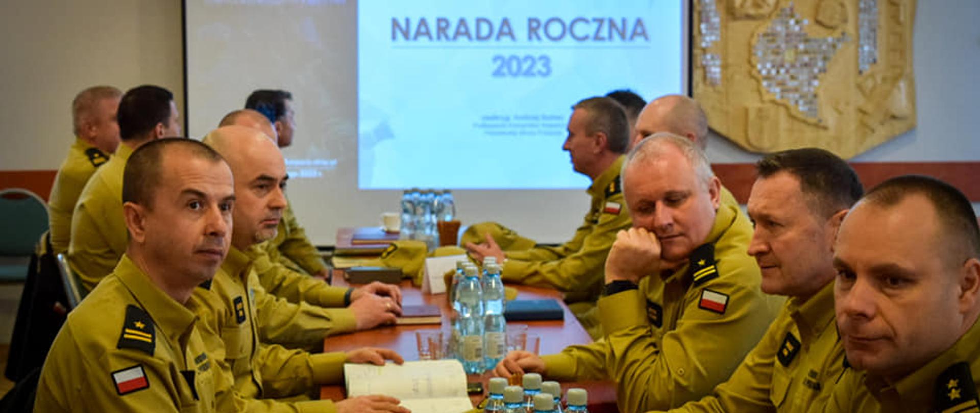 Kilkunastu mężczyzn w strażackim umundurowaniu, siedzących po dwóch stronach stołu. W tle ekran na którym wyświetlany jest slajd z namisem "Narada roczna 2023"