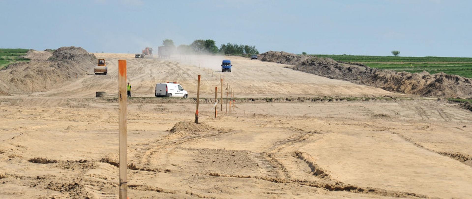 Plac budowy drogi ekspresowej - równy, szeroki pas piachu - w dużej odległości pojazdy i maszyny budowlane 