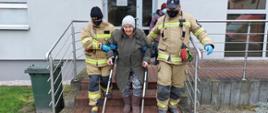 Dwoje strażaków zabezpieczonych w jednorazowe rękawiczki i maseczki pomaga zejść ze schodów starszej kobiecie poruszającej się przy pomocy kul ortopedycznych. Strażacy podtrzymują kobietę pod ręce i tak jej pomagają zejść. 
