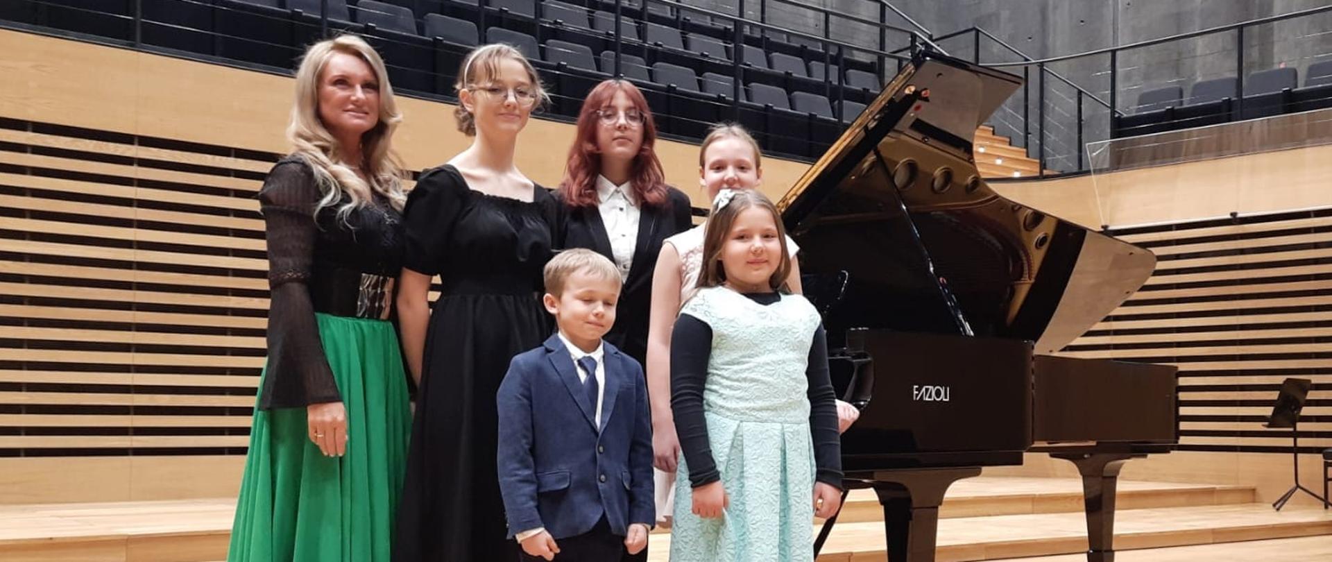 Pani Iwona Ulbrich z czterema uczennicami i uczniem przy fortepianie na scenie sali koncertowej PSM