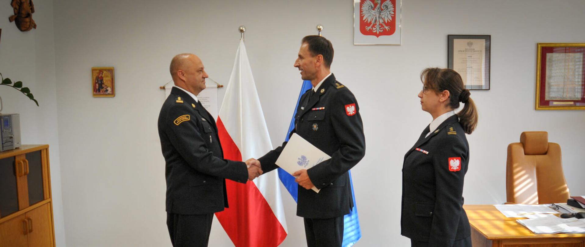 Dwóch mężczyzn w mundurach strażackich wita się uściskiem dłoni, a jeden z nich przekazuje teczkę. Obok nich stoi kobieta w mundurze, patrząca na nich. W tle widoczne są flagi Polski i Unii Europejskiej oraz godło Polski na ścianie