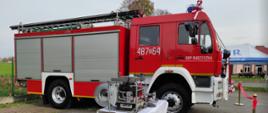 Zdjęcie przedstawia samochód ratowniczo gaśniczy straży pożarnej oraz sprzęt hydrauliczny ustawiony na stoliku. Przed samochodem wstęga koloru czerwonego