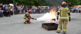 Park Słupna w Mysłowicach. Na placu odbywa się pokaz gaszenia pożaru przy użyciu gaśnicy. Na pierwszym planie dwóch strażaków w żóltych ubraniach specjalnych -Nomex i i chełmach. Jeden z nich trzyma w reku gaśnicę, która gasi pożar cieczy rozlanej na zbiorniku/tacy. Drugi stoi obok. W tle widzowie pokazu, stojący za barierkami. 