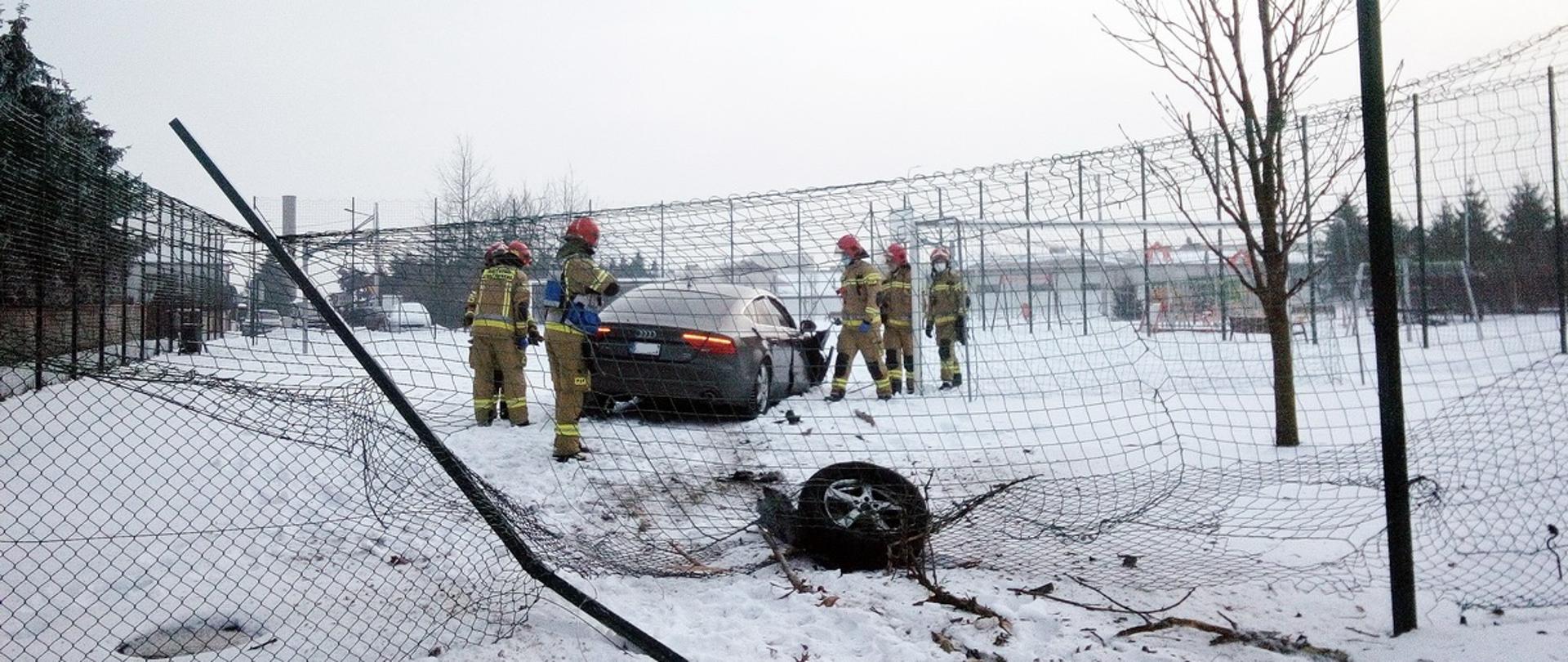 Zdarzenie komunikacyjne, w którym kierowca samochodu marki Audi pokonał ogrodzenie z siatki i wjechał na boisko. W koło samochodu znajdują się strażacy udzielając pomocy poszkodowanym oraz zabezpieczają miejsce zdarzenia. 