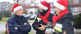 Korowód Wielkich Serc. Rawicz, ul Mikołajewicza. W okolicach siedziby ZUS stoi policjantka oraz dwie funkcjonariuszki PSP uczestniczące w akcji charytatywnej. Panie mają na sobie elementy stroju św. Mikołaja. W tle parking i budynki.