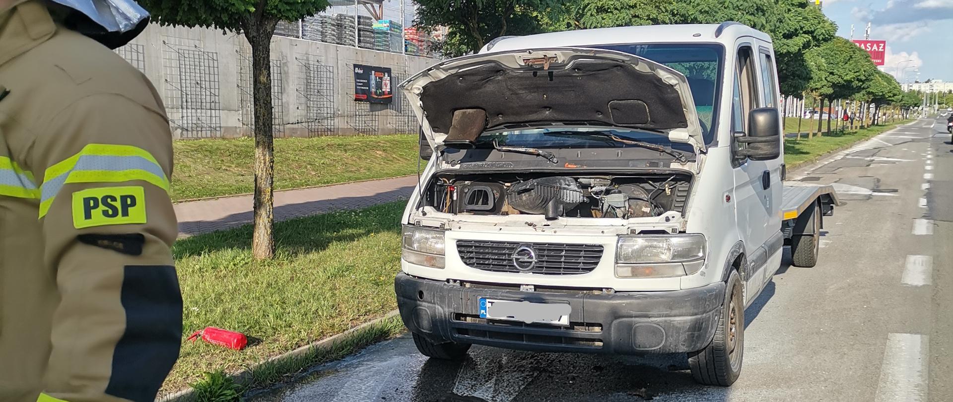Zdjęcie przedstawia Aleję Jerzego Szajnowicza-Iwanowa, przy której zaparkował samochód typu laweta gdzie doszło do pożaru w komorze silnika. Pojazd ma otwarta klapę silnika, na pierwszym planie widzimy strażaka a na ziemi leży wąż z prądownicą szybkiego natarcia.