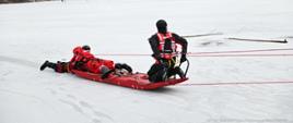 Strażacy ciągną po śniegu czerwone sanie lodowe. Mają na sobie kamizelki wypornościowe, a głowach mają kaski. W tle drzewa oraz jezioro pokryte lodem i śniegiem. Do deski przyczepione są liny.
