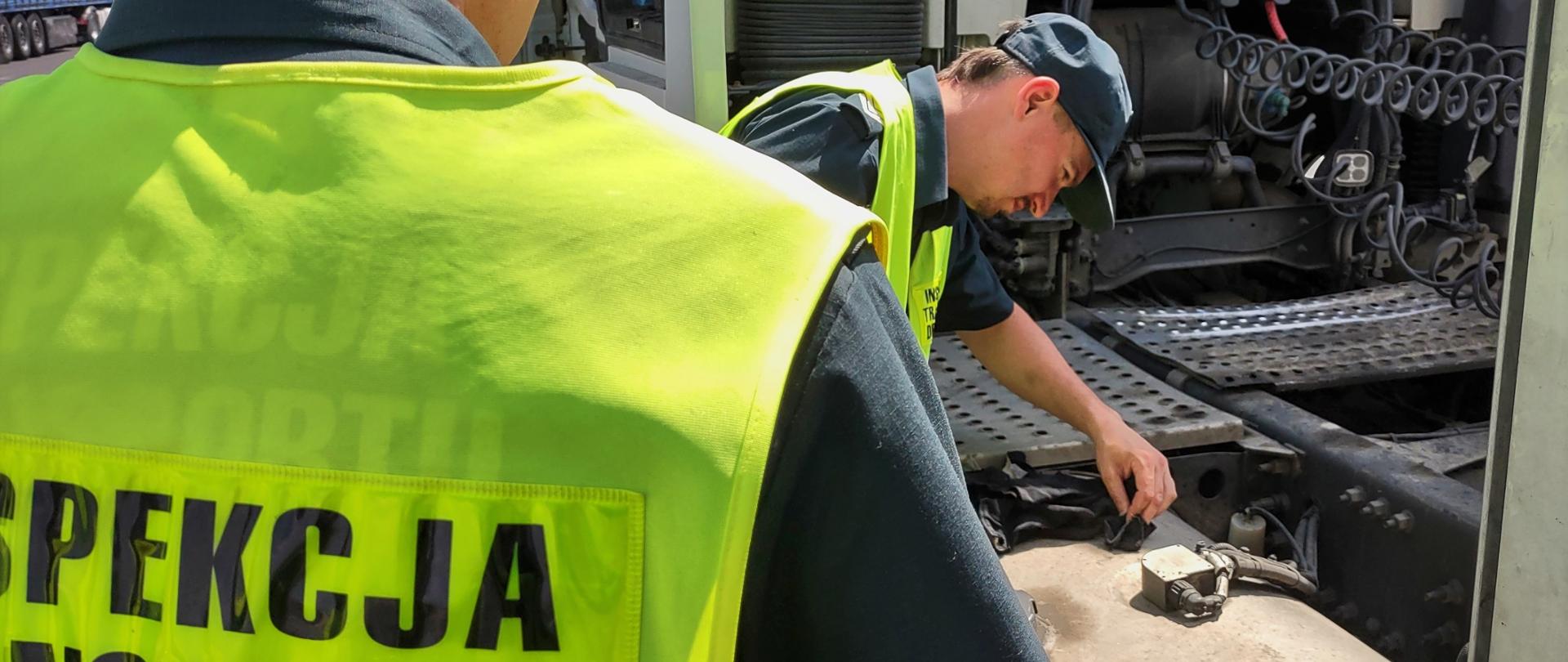 Dwóch inspektorów wielkopolskiej Inspekcji Transportu Drogowego stoi obok zatrzymanej ciężarówki i kontroluje pojemność zbiorników paliwa.
