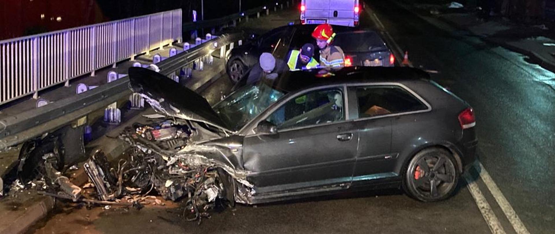 Na zdjęciu widać samochód marki Audi który dostawiony jest do barierek na moście i ma uszkodzony cały przód. Przy samochodzie stoi policjant, strażak i osoba w czapce. W tle samochód osobowy oraz radiowóz.