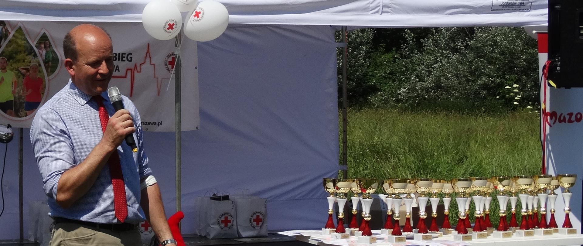 Na zdjęciu znajduje się mężczyzna mówiący do mikrofonu, po prawej stronie znajdują się upominki i puchary 