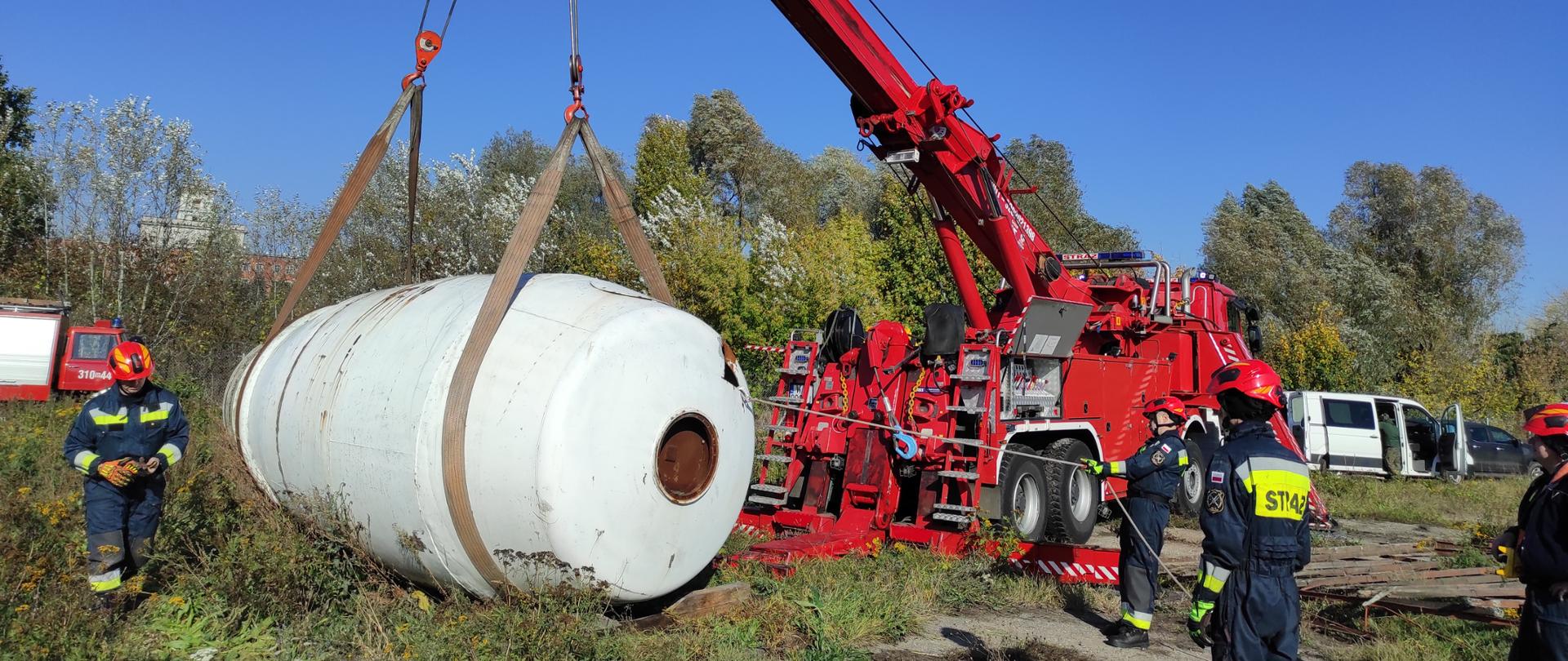 Zdjęcie przedstawia duży, czerwony żuraw ratowniczy zamontowany na ciężarówce, który unosi w powietrzu duży, owalny, biały obiekt przypominający zbiorni, przy użyciu pomarańczowych pasów nośnych.
Akcja ma miejsce w słoneczny dzień, na terenie trawiastym z widocznymi z tyłu drzewami o jesiennych barwach. W tle, po prawej stronie, widać zaparkowany biały samochód dostawczy.
Wokół czerwonej ciężarówki-żurawia, w centrum kadru, stoi trzech strażaków (lub ratowników technicznych) w czerwonych hełmach oraz specjalistycznym umundurowaniu, którzy nadzorują operację podnoszenia ładunku.
Obiekt podnoszony żurawiem jest koloru białego, cylindryczny z zaokrąglonymi końcami i ma ciemny, okrągły otwór w widocznej części.