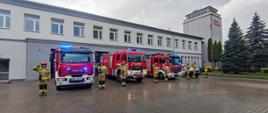Samochody strażackie wystawione przed budynek strażnicy z włączoną sygnalizacją świetlno - dźwiękową, funkcjonariusze ustawieni obok samochodów salutują na baczność