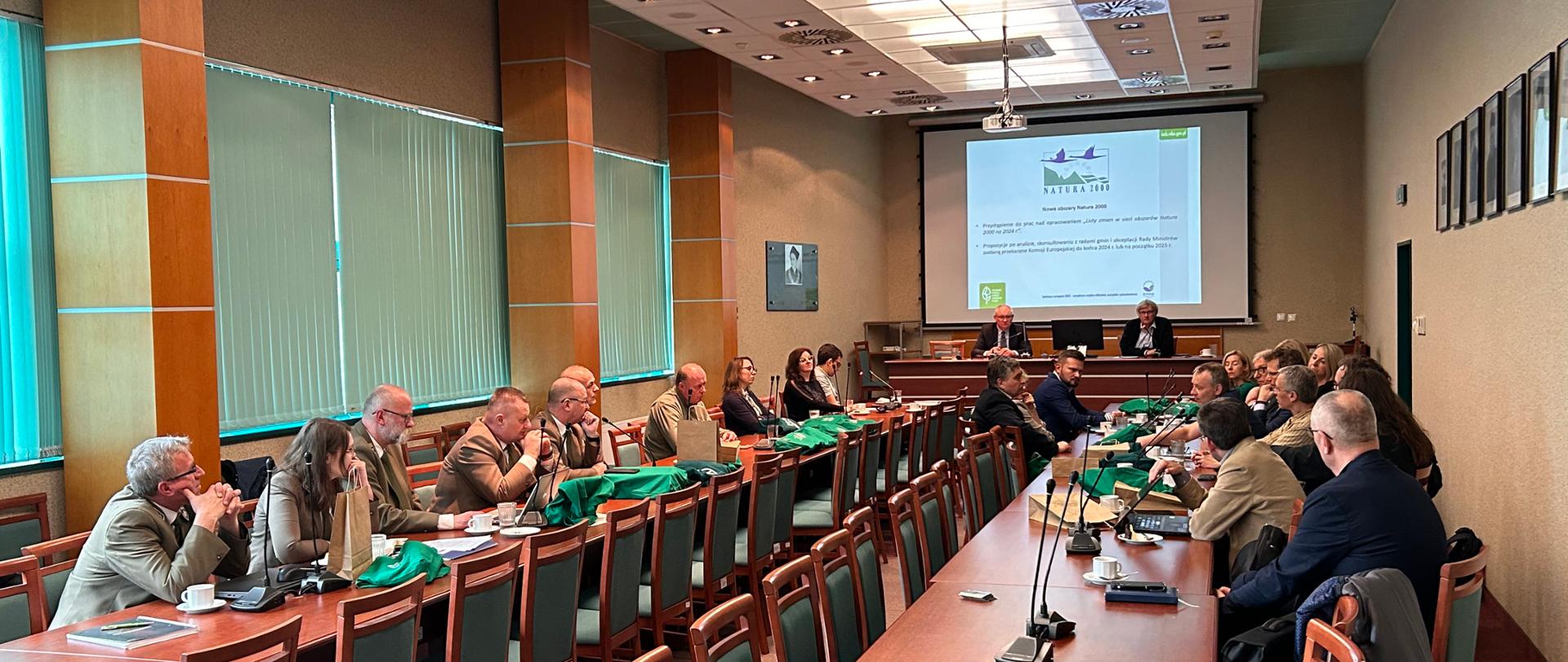 Sala, dwa długie stoły przy których siedzą ludzie. Na stołach mikrofony. W oddali na ekranie widoczna prezentacja, dwie osoby siedzą tyłem do ekranu, przodem do ludzi na sali. 