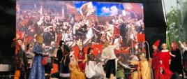 Zdjęcie przedstawia grupę osób ubranych w historyczne stroje, występujących na scenie. Za nimi znajduje się duży ekran wyświetlający obraz Jana Matejki „Hołd pruski” przedstawiający rozbudowaną scenę historyczną z wieloma postaciami zaangażowanymi w różne czynności. Wykonawcy odtwarzają scenę z malowidła.