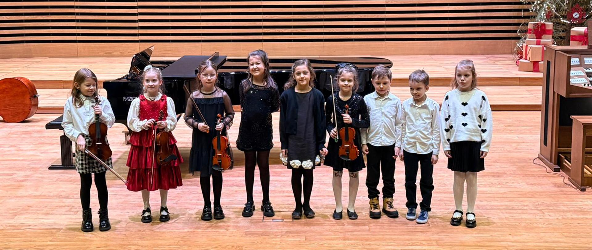 Siedem dziewczynek i dwóch chłopców stoi na scenie, cztery dziewczynki trzymają skrzypce.