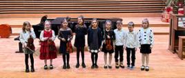 Siedem dziewczynek i dwóch chłopców stoi na scenie, cztery dziewczynki trzymają skrzypce.