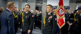 Generałowie PSP w mundurach galowych stoi naprzeciw ministra, który zwraca się do nich. Za nimi znajduje się strażak trzymający sztandar z orłem białym i hasłem „Bogu, Ojczyźnie”. W tle zgromadzeni funkcjonariusze i goście.