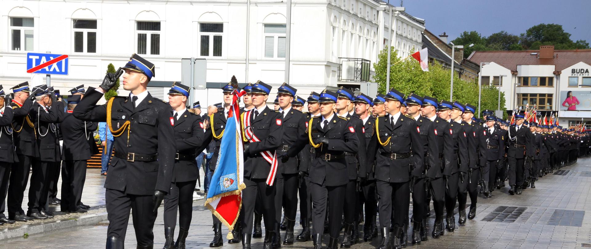 Grupa strażaków maszeruje. Strażak idący jako pierwszy salutuje. W tle widać budynki