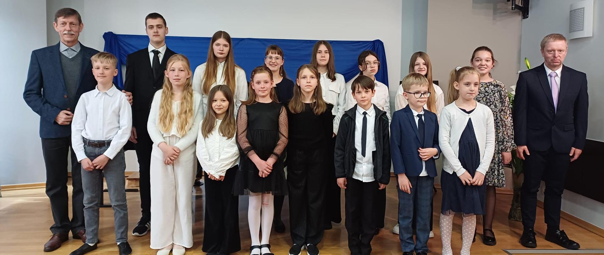 Zdjęcie wykonane podczas koncertu uczniów klasy fortepianu Pana Zbigniewa Kąkola i Pana Andrzeja Nowaka. Na zdjęciu grupa odświętnie ubranych uczniów, którzy wystąpili podczas koncertu. Wszyscy stoją na drewnianej scenie. Po bokach dwóch mężczyzna w ciemnych garniturach. Tło obrazka w odcieniach beżu.