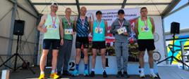 Grupa sześciu zawodników w medalach i z numerami startowymi stoi na podium pod zadaszoną sceną z banerem Szkoły Aspirantów PSP w Poznaniu; jeden z uczestników pokazuje kciuk w górę.