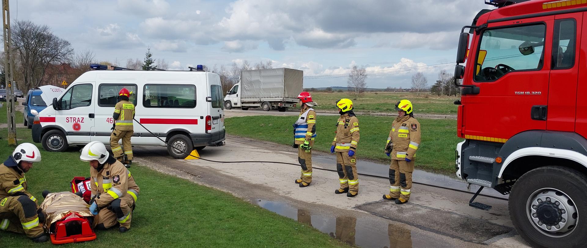 Strażacy udzielają pomocy medycznej poszkodowanemu kierowcy. W tle pojazd uczestniczący w zdarzeniu i samochód ratowniczo – gaśniczy.