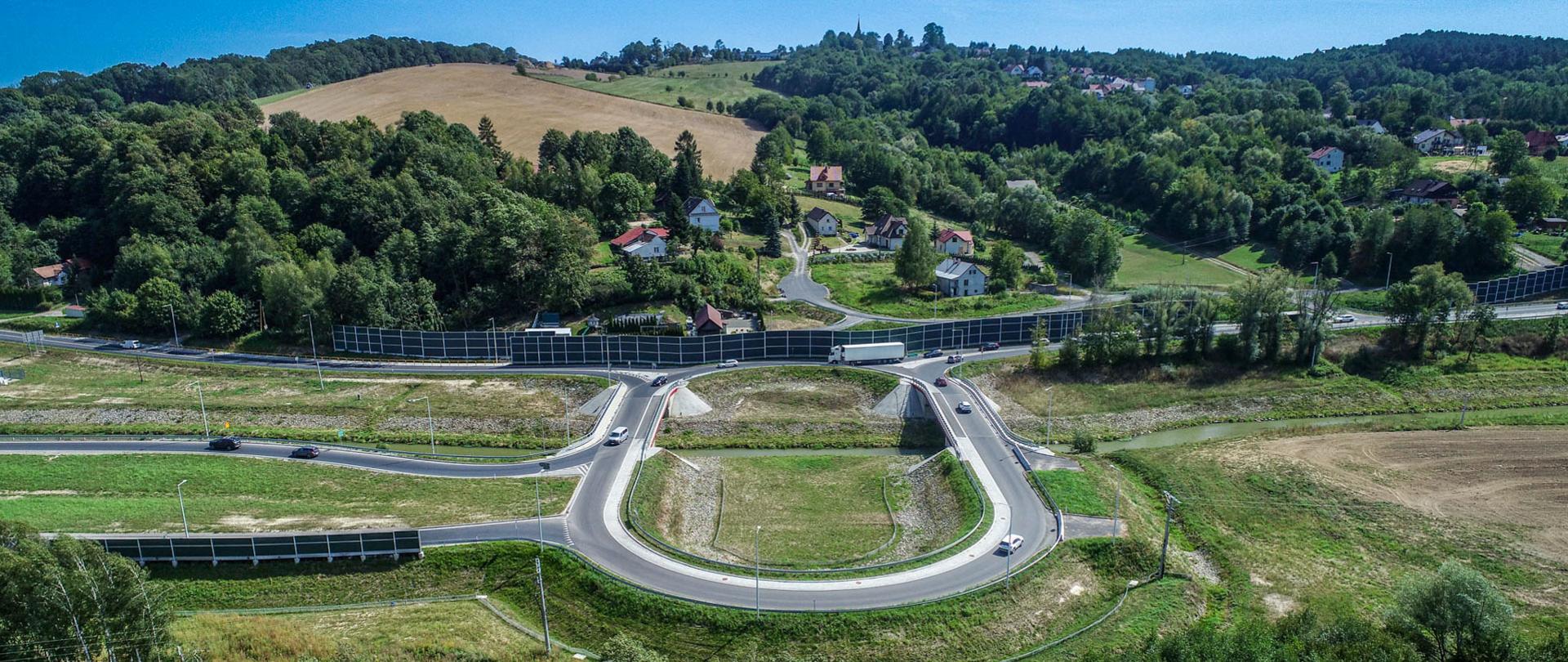 zdjęcie ronda w Okocimiu, po lewej zjazdy z ronda łączą się z drogą, po prawej prowadzą w kierunku pola, bezpośrednio za rondem zalesione w większości wzgórza i gdzieniegdzie zabudowania