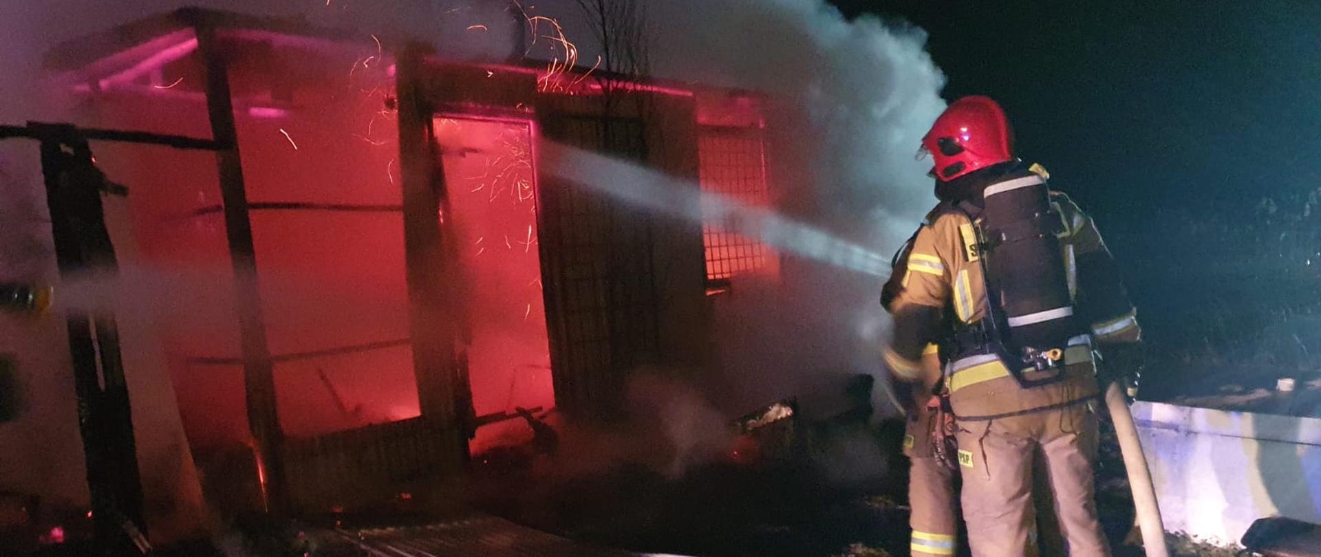 Na zdjęciu widać drewniany budynek objęty w całości pożarem oraz strażaków gaszących wodą 