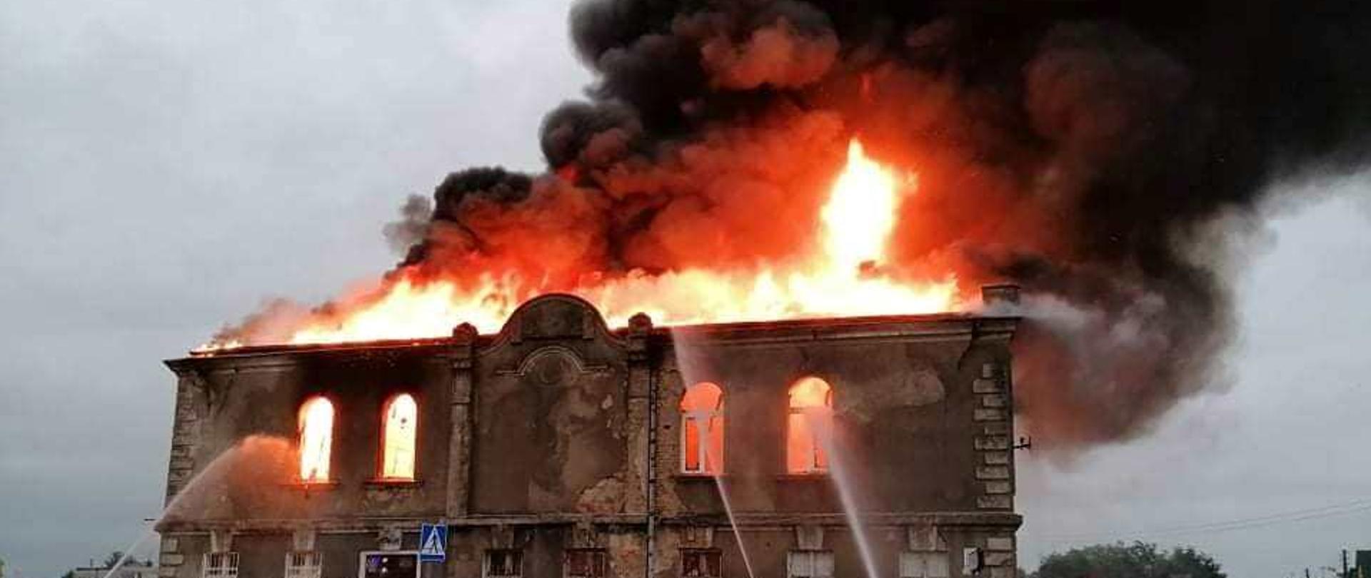 Z wnętrza budynku wydobywają się płomienie i dym, kilku strażaków gasi pożar prądami wody