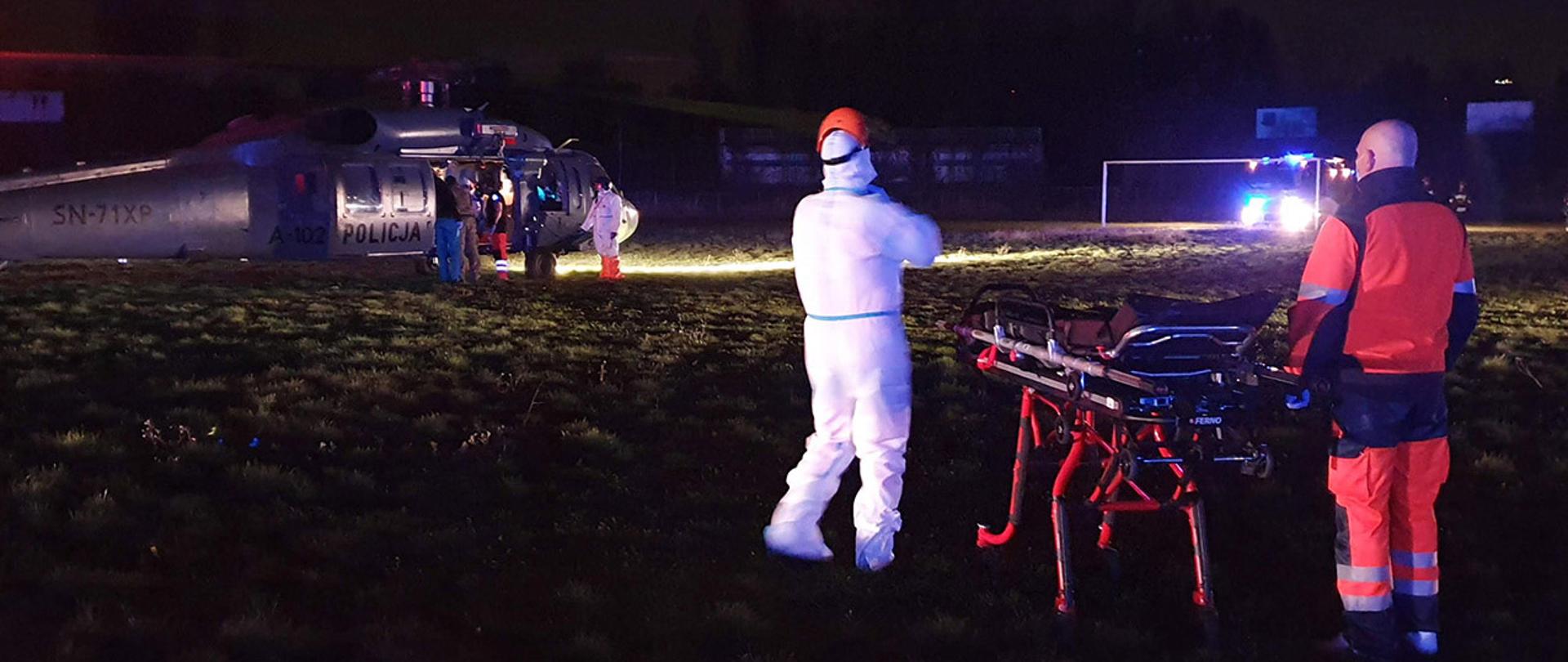 Na zdjęciu widać ratowników medycznych czekających z noszami na kółkach nocą na boisku trawiastym. Widać również stojący śmigłowiec i przy nim m.in. kolejnych ratowników. Boisko jest oświetlone jedynie reflektorami samochodu strażackiego.