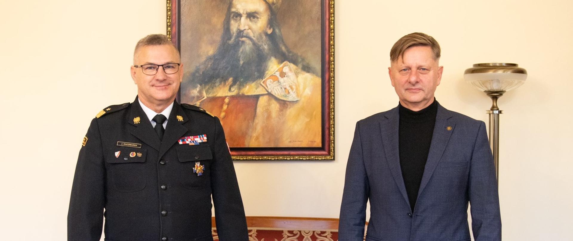 Komendant w mundurze wyjściowy, oraz rektor uczelni w pozycji stojącej. Za nimi portret Króla Kazimierza Wielkiego. 