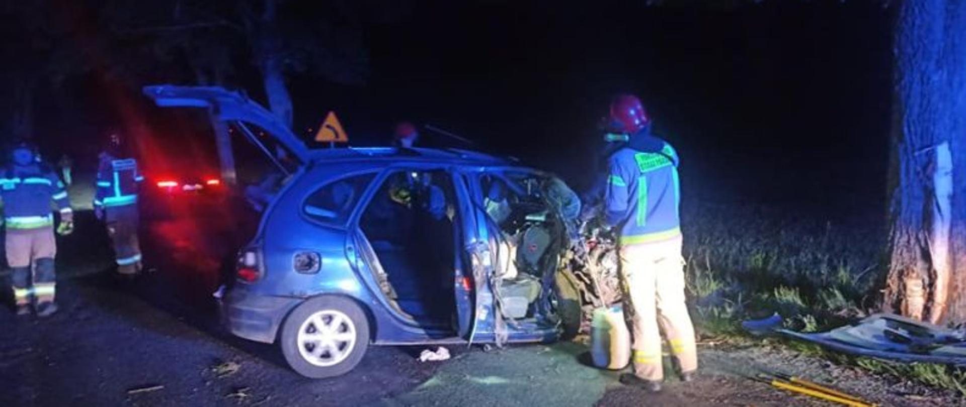 Centralny punkt zdjęcia stanowi rozbite auto. Auto stoi bokiem. Przy aucie jest strażak. Auto uderzyło w drzewo. Ma zniszczony całkowicie przód.