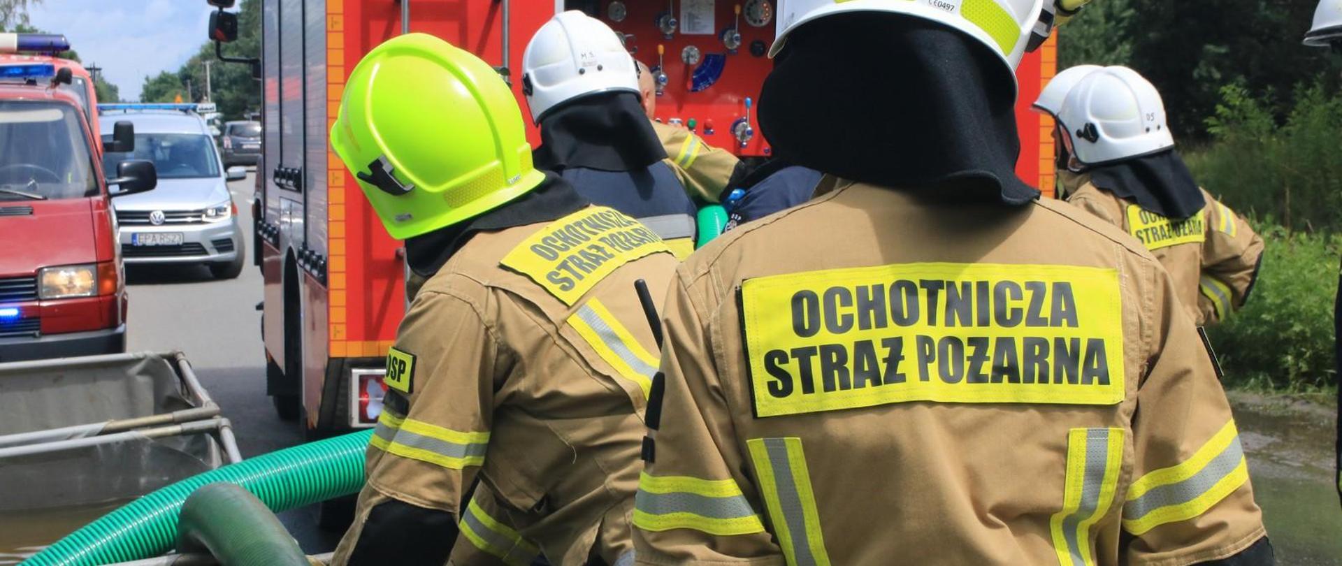 Na zdjęciu strażacy stoją przy brezentowych zbiornikach na wodę. Na drugim planie samochód strażacki pobierający z nich wodę.