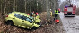 Po lewej stronie widać uszkodzony samochód osobowy znajdujący się w rowie. Za nim stoją strażacy i jedna osoba postronna. Po prawe stronie widać samochód pożarniczy blokujący drogę.