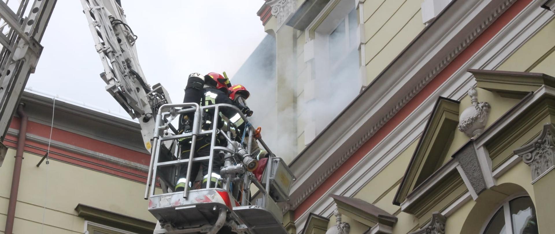 Na zdjęciu widać dwóch strażaków w koszu ratowniczym, ubranych w sprzęt ochrony układu oddechowego, którzy podjeżdżają pod okno starostwa powiatowego, z którego wydobywa się dym.