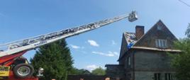 Na zdjęciu widać drabinę mechaniczną z rozłożoną drabiną, na której końcu znajduje się kosz z 2 strażakami. W oddali widać dom.