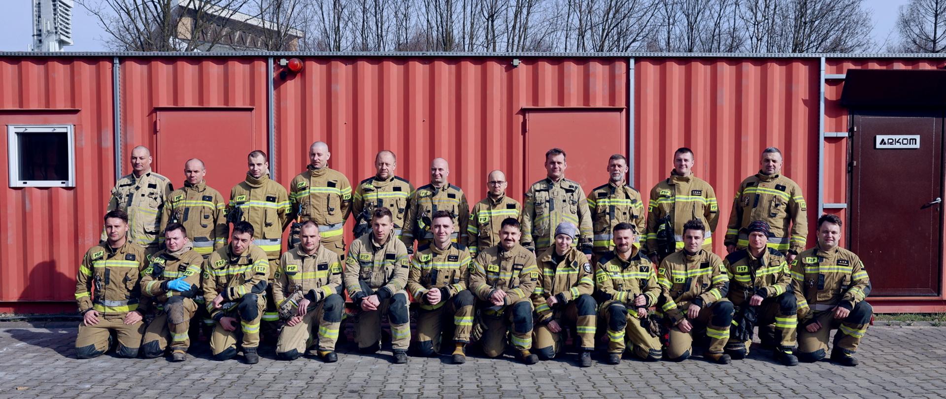 Pamiątkowe zdjęcie grupowe funkcjonariuszy PSP będących uczestnikami warsztatów dla instruktorów ogniowych z gaszenia pożarów wewnętrznych w towarzystwie strażaków będących instruktorami podczas warsztatów. Wszyscy ubrani w stroje bojowe pozują na tle trenażera